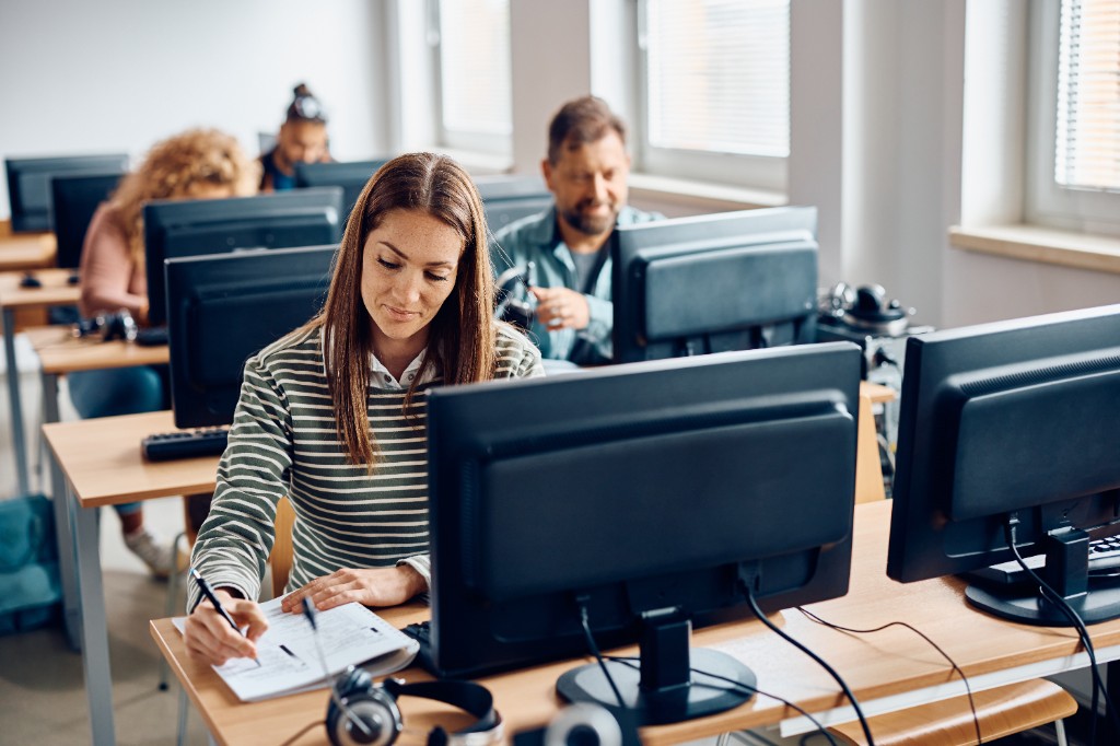 Student at a computer workstation in a classroom lab, with peers studying at nearby desks.